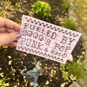 May include: A white piece of paper with a grid pattern and the text "Fueled by 2000's Pop Punk & Emo" written in black ink.