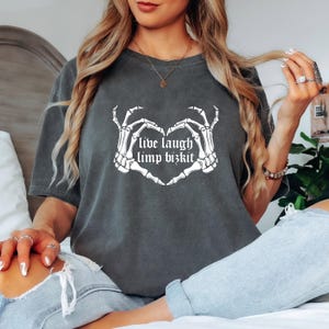 May include: Dark gray t-shirt with the text "Live Laugh Limp Bizkit" in a heart shape made of skeleton hands. The shirt is a casual style, suitable for everyday wear.