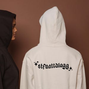 May include: Two hooded sweatshirts, one black and one cream-coloured, are shown from the back. The cream-coloured sweatshirt has the text "*stfuattalagg*" in a gothic font. The background is a solid brown.