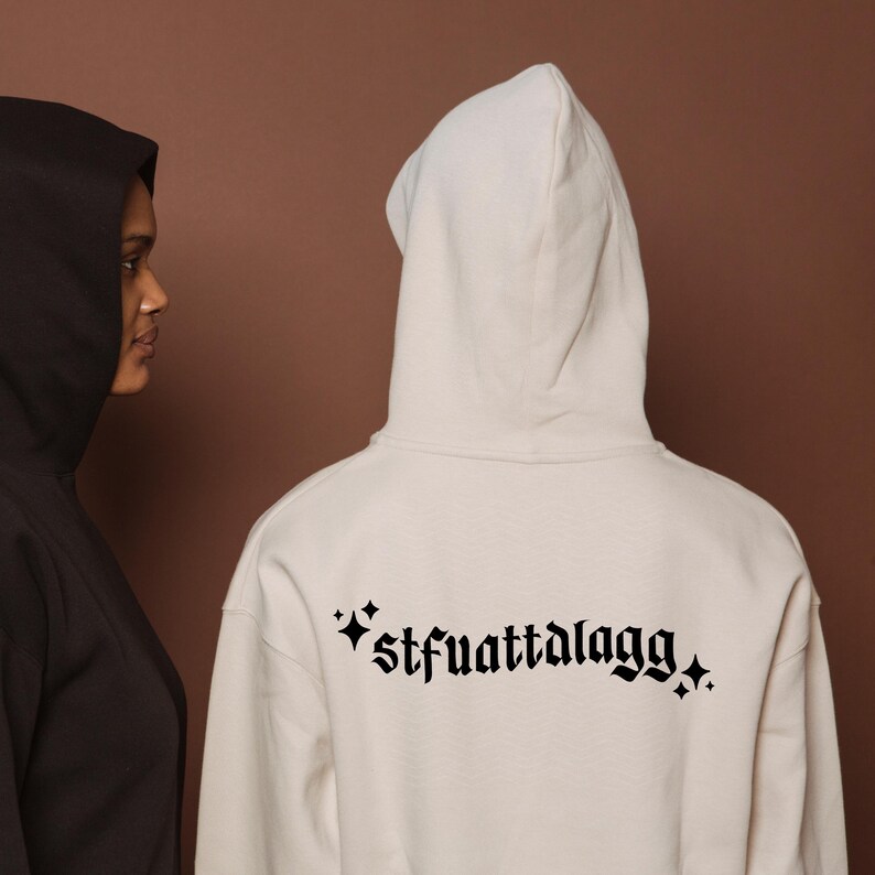 May include: Two hooded sweatshirts, one black and one cream-coloured, are shown from the back. The cream-coloured sweatshirt has the text "*stfuattalagg*" in a gothic font. The background is a solid brown.