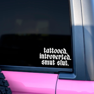 Op de afbeelding: Witte vinyl sticker met de woorden "tattooed. introverted. smut slut." in een gotisch lettertype. De sticker wordt weergegeven op een autoruit, met een roze carrosserie zichtbaar.