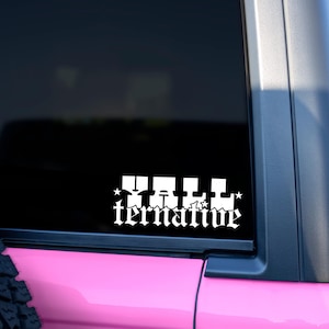 May include: White vinyl decal with the word "Tall" above "ternative" in a gothic font, with star accents. The decal is affixed to a vehicle window, with a pink body and black trim visible.