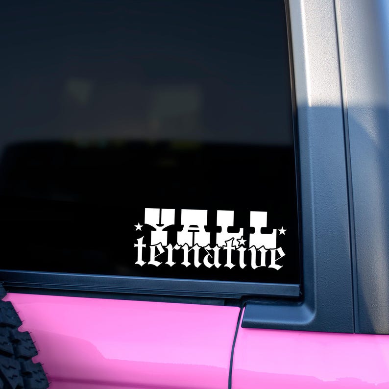 May include: White vinyl decal with the word "Tall" above "ternative" in a gothic font, with star accents. The decal is affixed to a vehicle window, with a pink body and black trim visible.