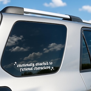 Könnte beinhalten: Weißer Vinyl-Aufkleber auf einer Autoscheibe mit dem Text "emotionally attached to fictional characters" in gotischer Schrift, mit Sternakzenten. Das Auto ist silberfarben und der Himmel ist blau mit Wolken.