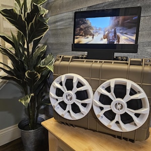 Peut inclure: Un système de divertissement portable de couleur beige avec un écran affichant un jeu vidéo. Deux haut-parleurs blancs avec un design complexe sont fixés au boîtier. Un pot de plante est visible en arrière-plan.