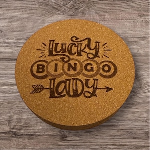 May include: A round cork coaster with the words "Lucky Bingo Lady" etched in a dark brown color. The coaster has an arrow pointing to the right and is sitting on a wooden surface.