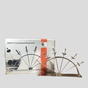 Puede incluir: Un juego de pinchos para fruta de plata con un soporte. El juego incluye siete pinchos con mangos decorativos en forma de espadas. La caja del juego es visible en el fondo y dice "WMF-Fruchtespießesgestell" y "WMF Tafelgerät".
