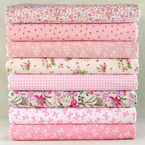 May include: A stack of eight folded fabric pieces in various pink floral, striped, and patterned designs. The fabrics include small flowers, stripes, gingham, and bow patterns. The fabrics are stacked neatly, showcasing a variety of textures and prints.