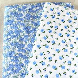 Puede incluir: Dos telas plegadas con estampados florales. Una tela presenta un patr&oacute;n denso de flores azules sobre fondo blanco. La otra tela tiene un patr&oacute;n de peque&ntilde;as flores azules con hojas verdes sobre fondo blanco. Las telas son probablemente de algod&oacute;n o lino.