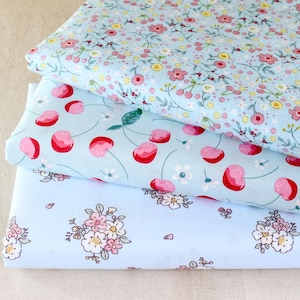Puede incluir: Tres pilas de tela con estampados florales y de frutas. La tela superior tiene un fondo azul claro con peque&ntilde;as flores rosas y amarillas. La tela del medio tiene un fondo azul claro con cerezas rojas. La tela inferior tiene un fondo azul claro con flores rosas.