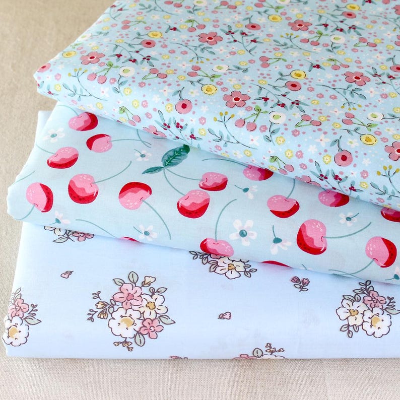 Puede incluir: Tres pilas de tela con estampados florales y de frutas. La tela superior tiene un fondo azul claro con peque&ntilde;as flores rosas y amarillas. La tela del medio tiene un fondo azul claro con cerezas rojas. La tela inferior tiene un fondo azul claro con flores rosas.