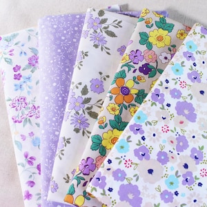 Puede incluir: Cinco muestras de tela con patrones florales en tonos de morado, amarillo y blanco. Las telas est&aacute;n dispuestas en una pila, con la muestra superior mostrando una gran flor amarilla con un centro morado.