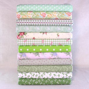 Puede incluir: Una pila de rollos de tela en tonos verdes y rosas. Las telas presentan patrones como corazones, diseños florales, cuadros vichy, lunares y lazos. Los rollos están apilados, mostrando una variedad de texturas y diseños.