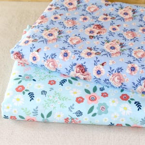 Puede incluir: Dos pilas de tela floral. La tela superior es azul claro con rosas rosas y blancas y detalles azules. La tela inferior es azul claro con margaritas, flores rojas y hojas verdes.