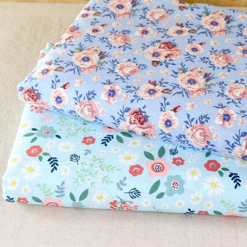 Puede incluir: Dos pilas de tela floral. La tela superior es azul claro con rosas rosas y blancas y detalles azules. La tela inferior es azul claro con margaritas, flores rojas y hojas verdes.