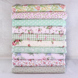 Può includere: Una pila di dieci pezzi di tessuto piegati con vari disegni floreali e fantasia. I tessuti presentano una gamma di colori, tra cui rosa, verde e bianco, con motivi come cuori, quadretti e righe. I tessuti sono impilati su una superficie chiara.