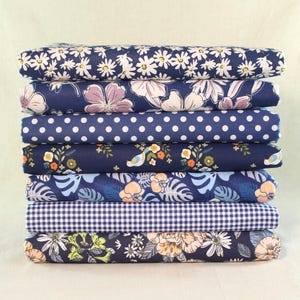 Puede incluir: Una pila de tela doblada con varios diseños florales y estampados. Las telas presentan margaritas blancas, flores moradas y blancas, lunares blancos, pájaros y hojas tropicales sobre un fondo azul marino. También hay una tela de cuadros azul y blanco.