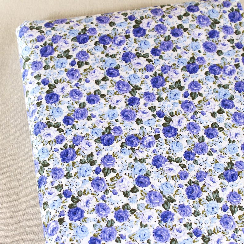 Puede incluir: Un rollo de tela con un estampado floral. El dise&ntilde;o presenta una densa disposici&oacute;n de rosas azules y moradas con hojas verdes sobre un fondo blanco. La tela est&aacute; enrollada, mostrando el motivo repetitivo.