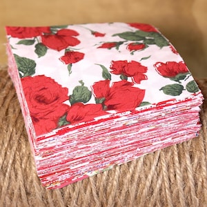 May include: A stack of square paper napkins with a floral rose pattern. The napkins feature large red roses with green leaves on a white background. The edges of the napkins are red and white.