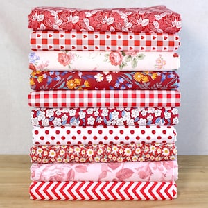 Może przedstawiać: Stos dziesięciu złożonych kawałków materiału w różnych czerwono-białych wzorach. Wzory obejmują kwiaty, kratę, kropki i zygzaki. Tkaniny są ułożone na drewnianej powierzchni.