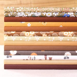 Può includere: Una pila di pezzi di tessuto in vari colori e motivi. I tessuti includono disegni floreali, a foglie e geometrici in tonalità di marrone, giallo e grigio. I tessuti sono ordinatamente piegati e impilati, mostrando la varietà di trame e stampe.