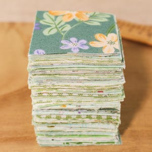 Peut inclure: Une pile de petits carrés de tissu avec un motif floral. Le tissu a un fond vert d'eau avec des fleurs orange, violettes et blanches et des feuilles vertes. La pile est posée sur une surface en bois.