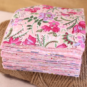 Może przedstawiać: Stos różowych kwadratów materiału w kwiaty. Tkanina ma powtarzający się wzór różowych i fioletowych kwiatów z zielonymi liśćmi na jasnoróżowym tle. Tkanina jest ułożona na rolce brązowego sznurka.
