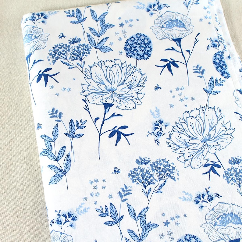 Puede incluir: Tela blanca con un estampado floral azul. El dise&ntilde;o presenta flores grandes y detalladas, hojas y grupos florales m&aacute;s peque&ntilde;os en varios tonos de azul. La tela est&aacute; doblada, mostrando la repetici&oacute;n del patr&oacute;n.