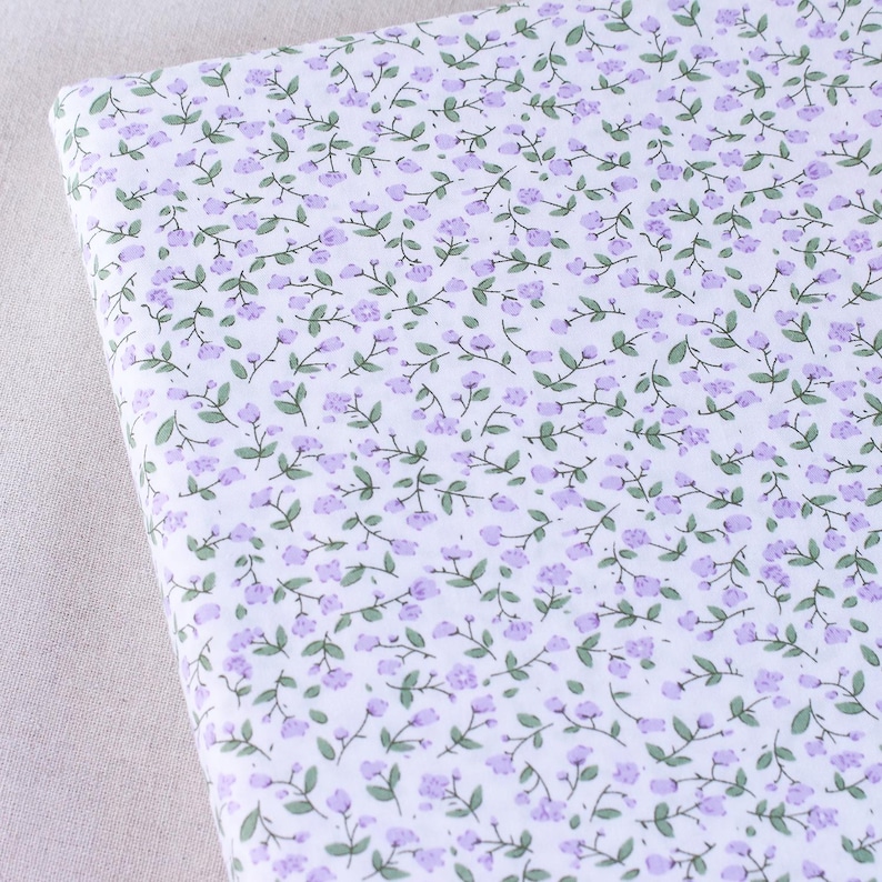Puede incluir: Tela blanca con un peque&ntilde;o estampado floral morado. Las flores est&aacute;n dispuestas en un patr&oacute;n repetitivo en la tela.