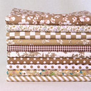 Puede incluir: Una pila de varias piezas de tela estampada en tonos marrones, beige y blanco. Los estampados incluyen flores, lunares, cuadros vichy y rayas. Las telas están cuidadosamente dobladas y apiladas, mostrando una variedad de diseños.