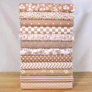 Könnte beinhalten: Ein Stapel gerollter Stoffballen in verschiedenen Mustern und Farben. Die Stoffe weisen Blumen-, Tupfen-, Herz- und geometrische Designs in den Farbtönen Beige, Weiß und Braun auf. Die Stoffe sind auf einer Holzoberfläche gestapelt.