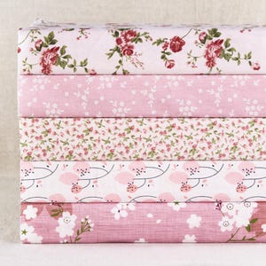 Pode incluir: Uma pilha de pedaços de tecido com vários padrões florais. Os tecidos apresentam fundos rosa e branco com desenhos de rosas vermelhas, pequenas flores brancas e flores de cerejeira rosa. Ideal para projetos de costura.
