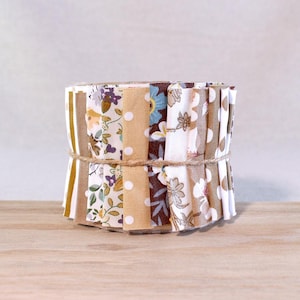 May include: A fabric jelly roll bundle featuring a variety of strips with floral, polka dot, and solid patterns in shades of brown, tan, white, and blue, all secured together with a thin piece of twine wrapped around the centre of the stack.