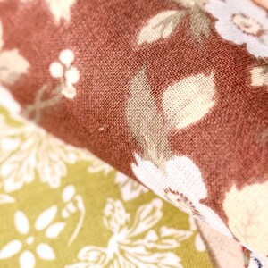 Pode incluir: Close-up de tecido floral em tons de castanho, creme e verde. O tecido tem uma apar&ecirc;ncia texturizada com um padr&atilde;o repetido de flores e folhas. A imagem destaca os detalhes do t&ecirc;xtil.