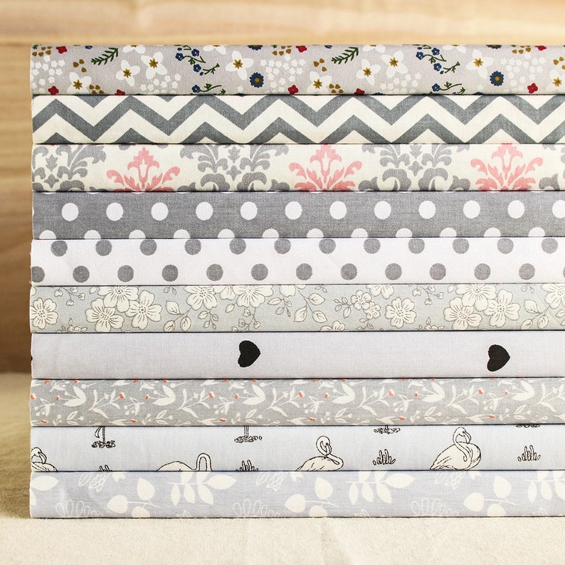 May include: A stack of 12 fabric swatches in various patterns and colors, including gray, white, pink, and floral prints. The fabrics are arranged in a neat stack, with the top fabric being a light gray with a floral pattern.