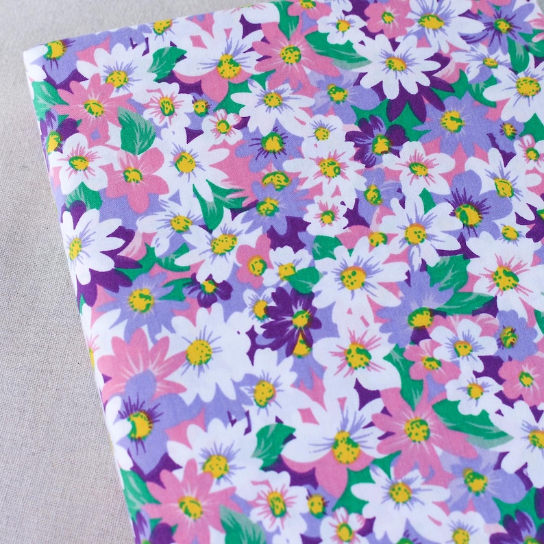 Puede incluir: Una tela blanca con un estampado floral con flores rosas, moradas y verdes. Las flores son de dise&ntilde;o de margarita con centros amarillos.