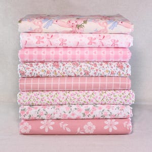 May include: A stack of pink fabric rolls with various patterns. Designs include floral, gingham, bows, and hearts. The fabrics are in shades of pink and white, suitable for sewing and crafting projects.