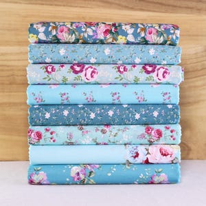 May include: A stack of eight fabric rolls, each displaying a different floral pattern. The fabrics feature a variety of blue hues, with pink, purple, and white flower designs. The rolls are neatly stacked, showcasing the diverse patterns.