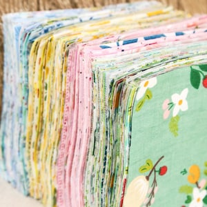 Może przedstawiać: Stos kolorowych kwadratów materiału w różnych wzorach i kolorach, w tym niebieskim, żółtym, różowym i zielonym. Tkaniny mają kwiatowe i geometryczne wzory, idealne do patchworku lub projektów rzemieślniczych.