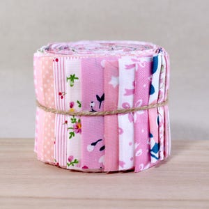 May include: A roll of pink and white fabric strips tied with twine. The fabric features various patterns, including polka dots, stripes, and floral designs. The color palette includes shades of pink and white, creating a soft and feminine aesthetic. The fabric is likely for quilting or crafting.