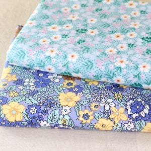 Puede incluir: Dos piezas de tela dobladas con estampados florales. La tela superior es azul claro con flores blancas y rosas y hojas verdes. La tela inferior es morada con flores amarillas, azules y blancas y hojas verdes. Las telas son probablemente de algod&oacute;n o lino.