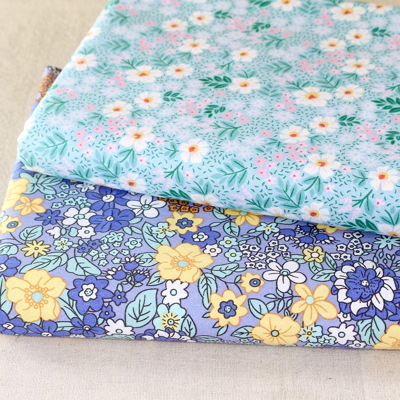 Puede incluir: Dos piezas de tela dobladas con estampados florales. La tela superior es azul claro con flores blancas y rosas y hojas verdes. La tela inferior es morada con flores amarillas, azules y blancas y hojas verdes. Las telas son probablemente de algod&oacute;n o lino.