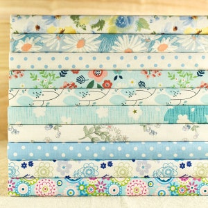 May include: A stack of fabric squares with various floral and geometric patterns in shades of blue, white, and pink. The fabric squares are arranged in a vertical stack, with the top square being a light blue floral pattern and the bottom square being a colorful geometric pattern.