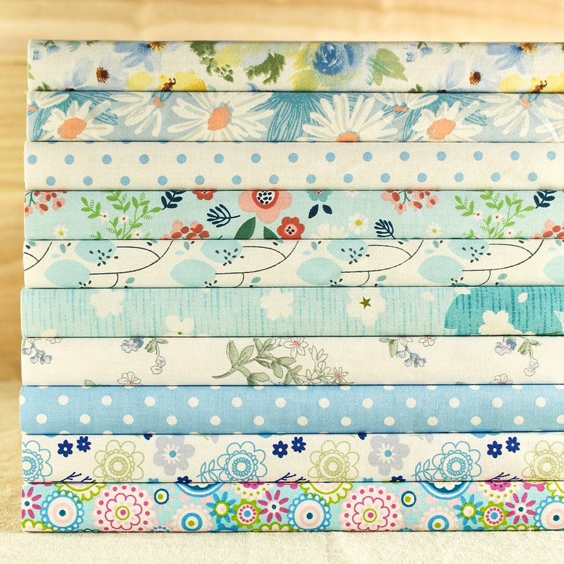 May include: A stack of fabric squares with various floral and geometric patterns in shades of blue, white, and pink. The fabric squares are arranged in a vertical stack, with the top square being a light blue floral pattern and the bottom square being a colorful geometric pattern.