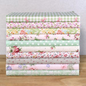 Könnte beinhalten: Ein Stapel gemusterter Stoffe in verschiedenen Designs und Farben. Die Stoffe umfassen Blumen-, Vichy- und Streifenmuster in Rosa-, Grün- und Weißtönen. Die Stoffe sind ordentlich auf einer Holzoberfläche gestapelt.