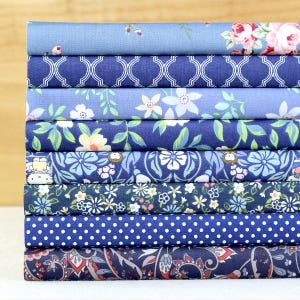 May include: A stack of various patterned fabric rolls. The fabrics feature floral, geometric, and polka dot designs in shades of blue and white. The top roll has a light blue background with pink flowers.