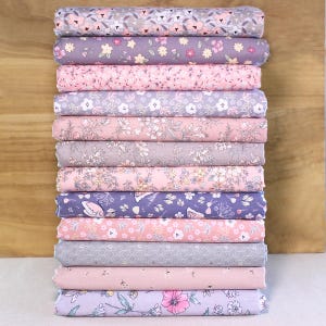 May include: A stack of floral fabric rolls in various pastel colors, including pink, purple, and gray. Each roll displays a different floral pattern, creating a visually appealing display. The fabric is likely cotton or a similar material, suitable for quilting or sewing projects.