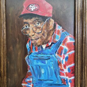May include: Oil painting portrait of a person wearing a red baseball cap with a white and red logo, glasses, and a blue and red plaid shirt with blue overalls. The artwork is framed in a brown wooden frame.