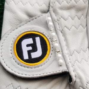 May include: White leather golf glove with a black and yellow FJ logo and a row of white pearls on the cuff.