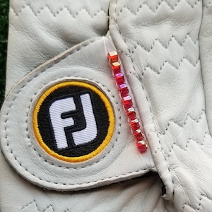 May include: White golf glove with the FootJoy logo in black, yellow, and white. The glove has a decorative line of red and orange rhinestones. The glove is made of leather and has a quilted design on the back.